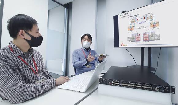 ▲SK텔레콤은 100Gbps 전송기술 기반의 암호 기술로 한국암호모듈검증(KCMVP) 인증을 획득했다고 밝혔다. 사진은 SK텔레콤 직원들이 KCMVP 인증 암호모듈 및 해당 모듈이 설치된 전송장비에 대해 논의하는 모습 / 사진=SK텔레콤